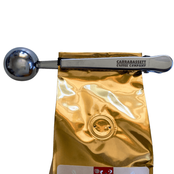Stainless steel coffee scoop with a clip on the handle end. The words: "Carrabassett Coffee Company" are embossed on the handle. The scoop is shown clipped to a gold one pound coffee bag in this image.