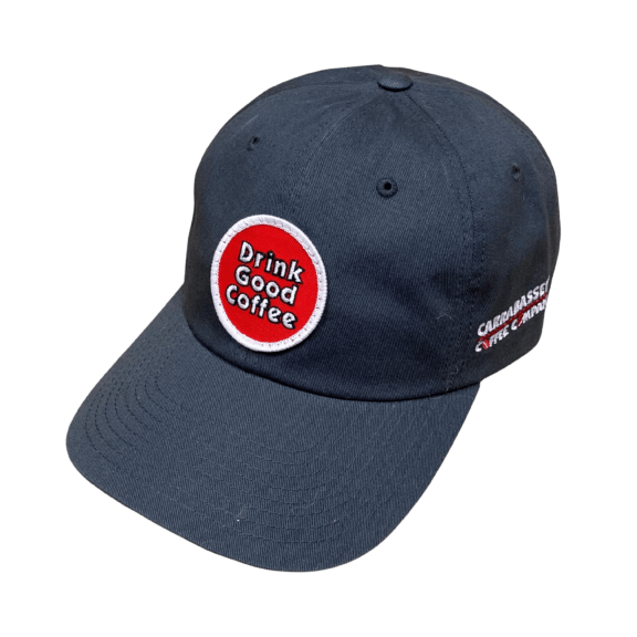 Charcoal gray unstructured baseball hat with curved brim. This hat features a circle embroidered patch in red with a white border. Text on the patch reads; Drink Good Coffee in white outlined in black.
