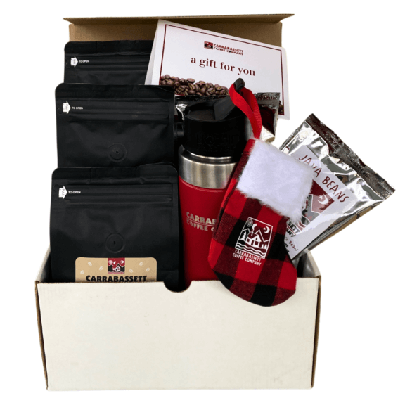 A white box containing all the items in the holiday bundle: 3 12 oz bags of Carrabassett Coffee, a gift for you card, two 4 oz bags of chocolate covered espresso beans, a red stainless steel travel mug, and a fabric red buffalo plaid stocking ornament with our logo on it.