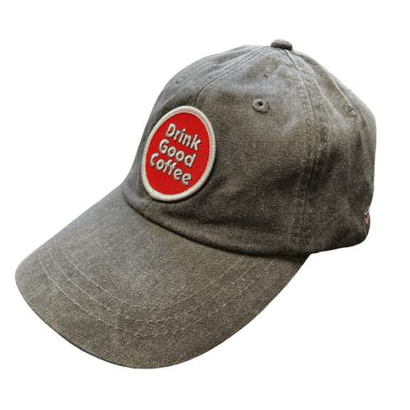 Gray unstructured baseball hat with curved brim. This hat features a circle embroidered patch in red with a white border. Text on the patch reads; Drink Good Coffee in white outlined in black.