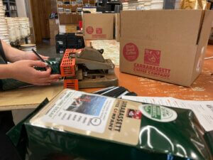 photo of a carrabassett coffee company worker using the heat sealer to seal a coffee bag. In the foreground, you can see an already sealed one pound bag of Carrabassett coffee (Organic Gondi Line Decaf), and Carrabassett coffee company shipping boxes. Preparing an order for shipment.