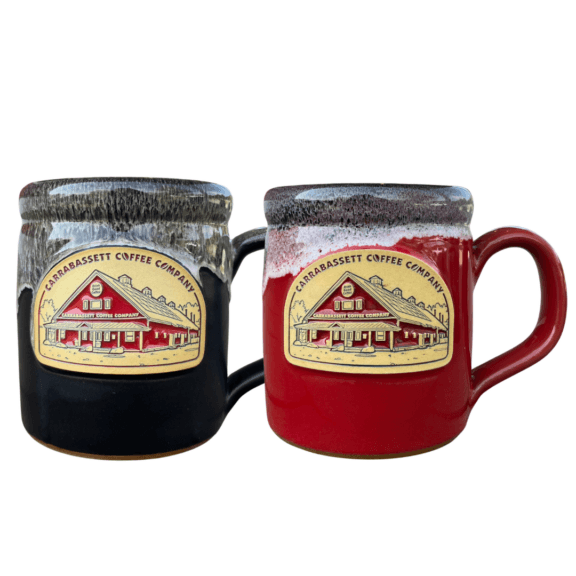 Two Carrabassett Coffee Ceramic Mugs. One Black with a sand marble drip. One Red with a black and white marble drip. Both feature a medallion with a red glazed Drawings of the Carrabassett coffee Company Roastery Building.